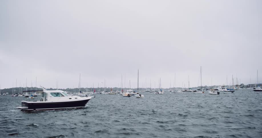 A motorboat slowly navigates through a choppy harbor filled with moored sailboats on an overcast day
