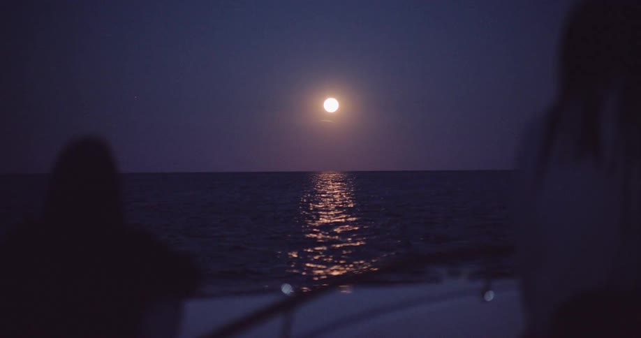 A serene view from a boat shows the full moon casting a shimmering reflection across the calm sea at night