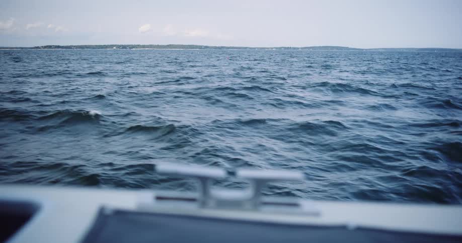 A slow-motion point-of-view shot from the bow of a boat sailing on the open ocean towards a distant shoreline