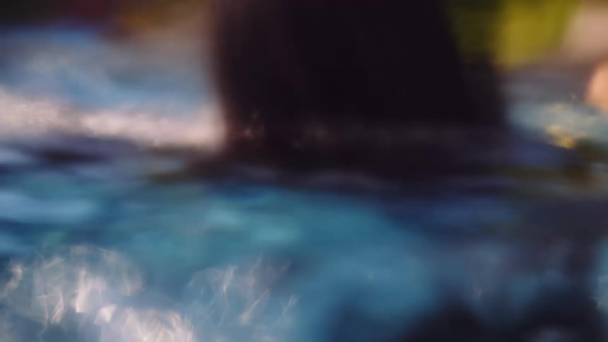 A person with dreadlocks wearing a necklace slowly emerges from underwater in a swimming pool in an artistic slow motion shot