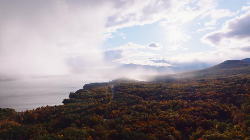 A breathtaking aerial view shows a vibrant autumn forest bordering a misty lake, with stunning sun rays breaking through the clouds over distant mountains