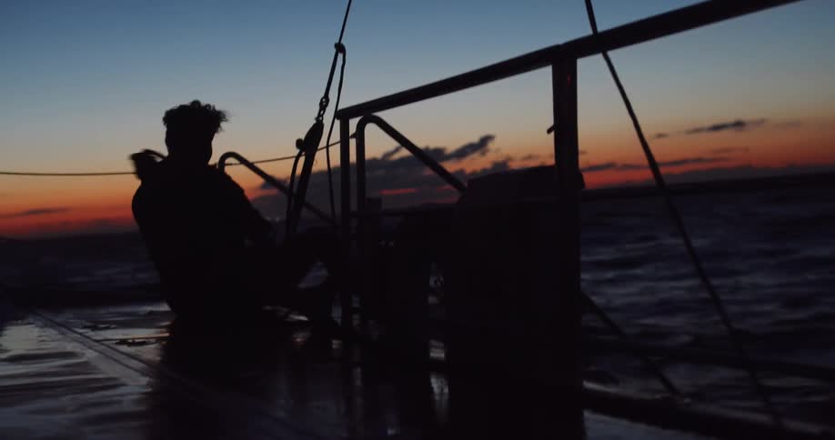 A silhouetted sailor sits on the deck of a rocking sailboat before standing up and walking away against a beautiful twilight sky over the ocean