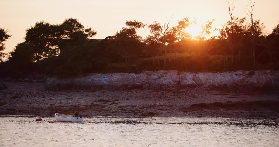 A small white motorboat is moored in a calm bay as the sun sets over a tree-lined, rocky shore
