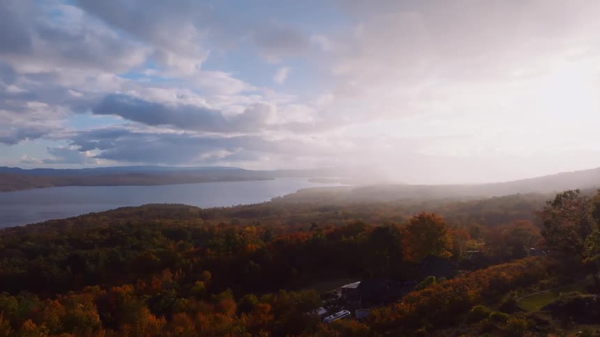 A breathtaking aerial view reveals a vast lake and colorful autumn forest under a hazy, sunlit sky