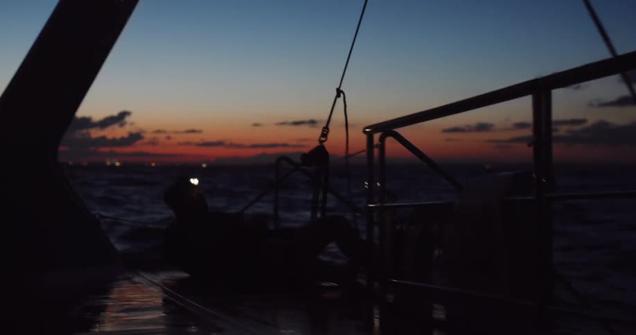 Sailor with a headlamp grinds a winch on the deck of a sailboat at dusk, silhouetted against the colorful sky