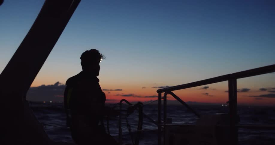 In silhouette against a beautiful twilight sky, a sailor with a headlamp works with ropes on the deck of a moving yacht
