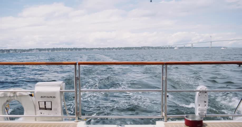 A scenic view from the stern of a boat as it cruises across the water, leaving a wake, with a distant bridge on the horizon under a cloudy sky