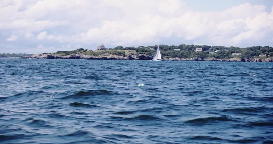 A scenic view from a boat on the choppy blue ocean shows a white sailboat passing a rocky, tree-lined coastline on a cloudy day
