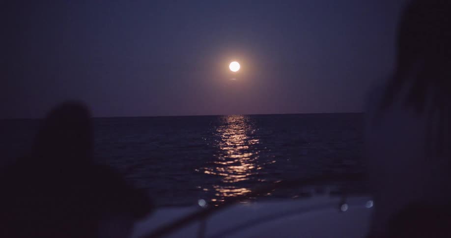 A serene full moon casts a beautiful reflection over the ocean as viewed from the back of a boat at night
