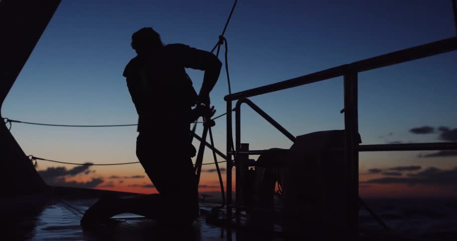 Silhouette of a sailor kneeling on the deck of a sailboat at twilight, securing ropes as the boat rocks on the ocean waves