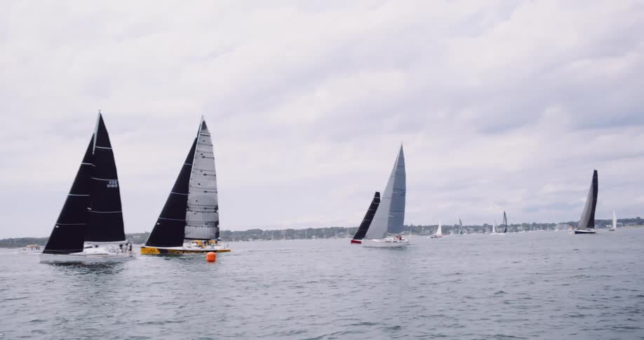 A fleet of modern sailboats with large sails competes in a regatta on the ocean under a cloudy sky