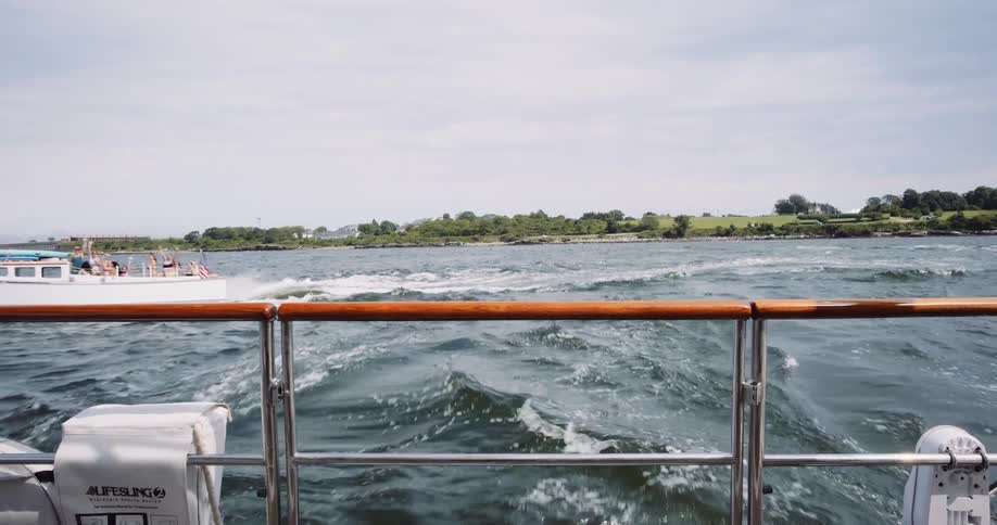 View from the stern of a boat as another launch with passengers passes by on a choppy sea near a green coastline