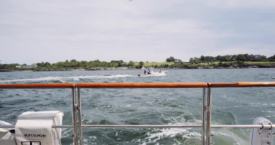 A serene slow-motion view from the back of a boat reveals its wake as another motorboat passes by with a lush shoreline in the background