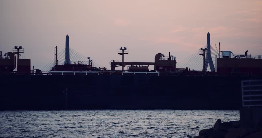 A large cargo ship and other boats sail through Boston Harbor at dusk, with the iconic Zakim Bridge silhouetted against the twilight sky