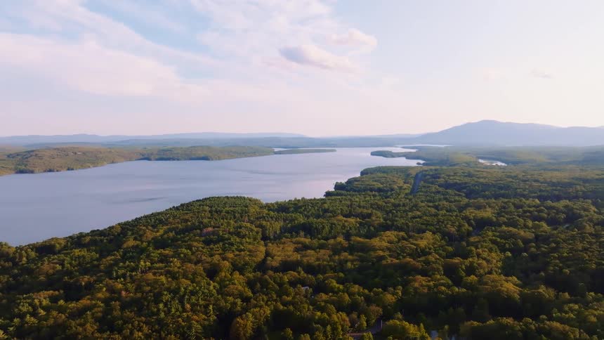 A static aerial shot shows a beautiful, expansive lake surrounded by a lush green forest with mountains in the background