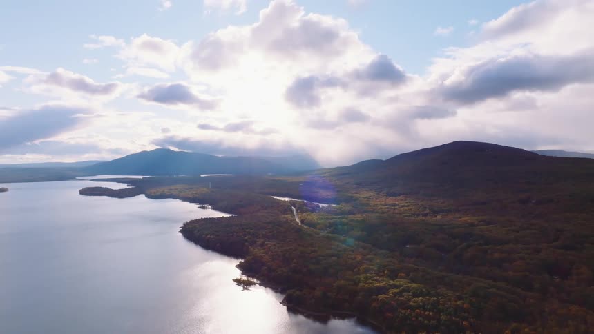A peaceful, static aerial view of a large mountain lake and rolling hills covered in dense autumn forests under a dramatic cloudy sky with sunbeams
