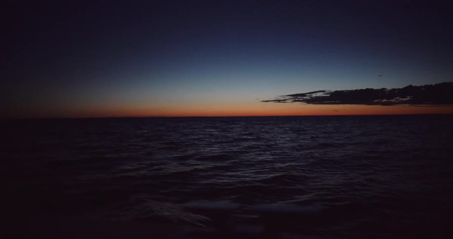 Gentle ocean waves move slowly as the sun sets, casting a faint orange glow on the dark horizon