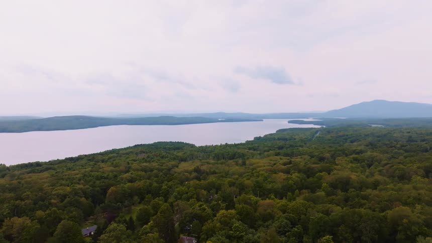 A serene aerial view of a vast lake surrounded by lush green forests and distant mountains on an overcast day