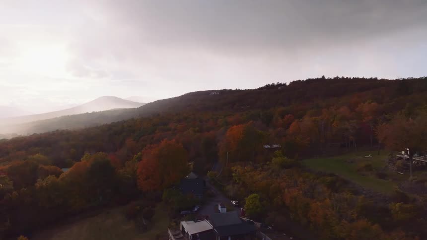 An expansive aerial view of homes on a mountainside reveals a vast forest of colorful autumn trees under a hazy morning sky