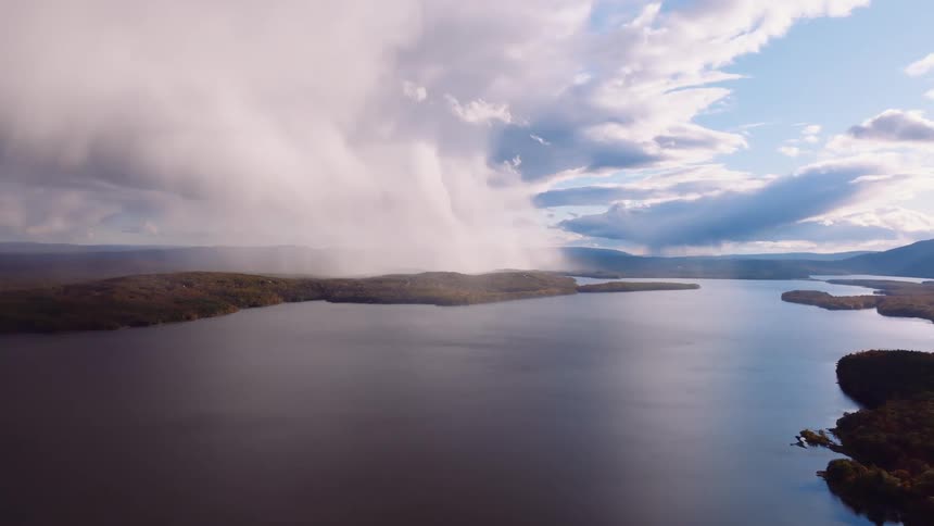 A stunning aerial view captures a powerful, localized rainstorm pouring down from dramatic clouds onto a vast lake surrounded by autumn forests