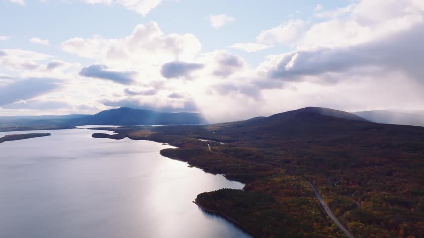 An epic aerial view of sun rays breaking through the clouds over a vast mountain lake and a colorful autumn forest