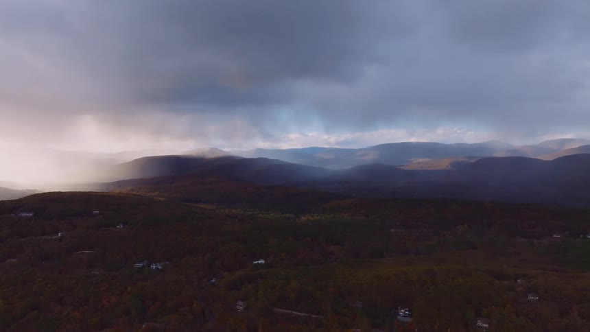 A dramatic aerial cinemagraph captures sunlight breaking through storm clouds over a vast mountain valley and autumn forest