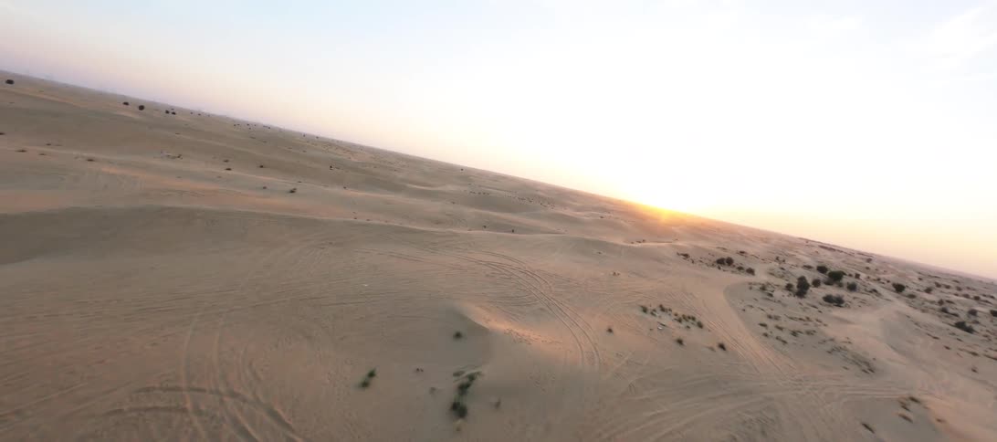 A dynamic FPV drone flies over the top of a unique modern building with a geometric roof, revealing the vast, beautiful desert dunes at sunrise