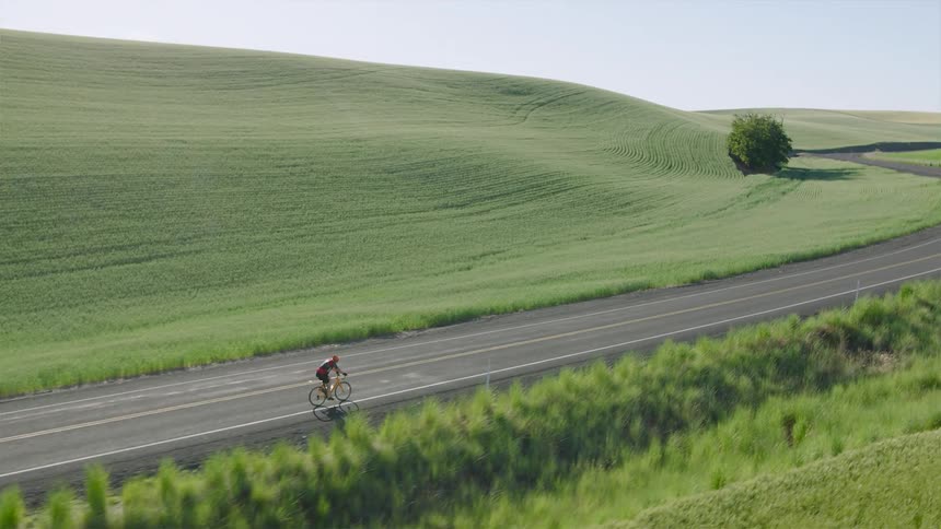 An aerial view shows a lone cyclist riding along a quiet country road surrounded by beautiful rolling green hills