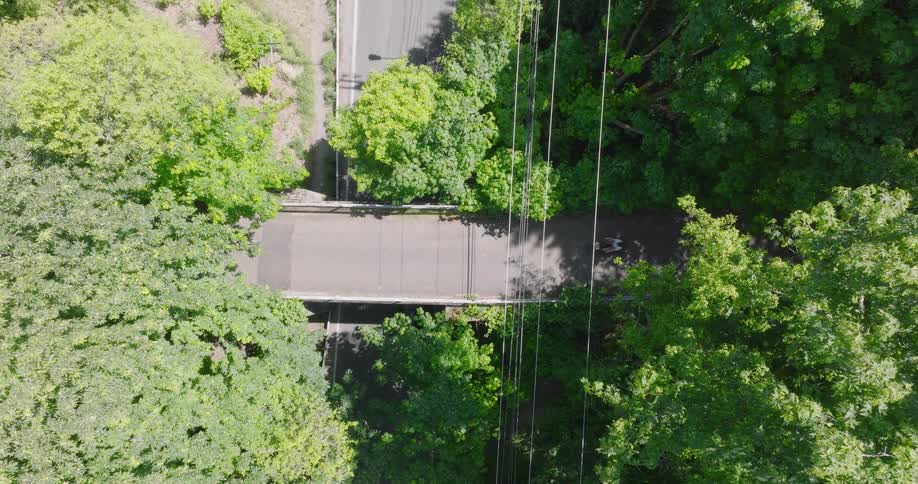 Top-down aerial shot captures cyclists passing under a bridge on a paved trail through a lush green forest