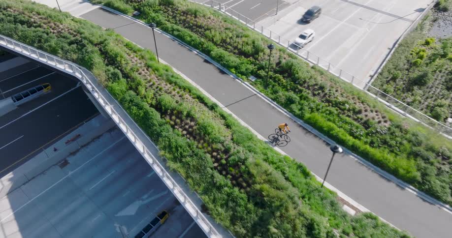 An aerial view follows a cyclist riding along a modern green roof bike path over a busy highway on a sunny day
