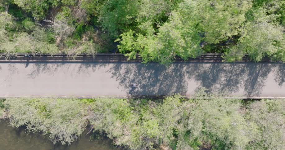 Top-down drone shot of a cyclist riding on a shaded path next to a river through a vibrant green forest