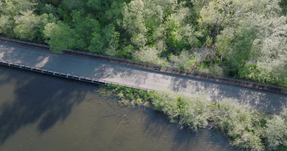 Top-down aerial view of a person cycling along a peaceful paved trail that runs over a river and alongside a lush green forest