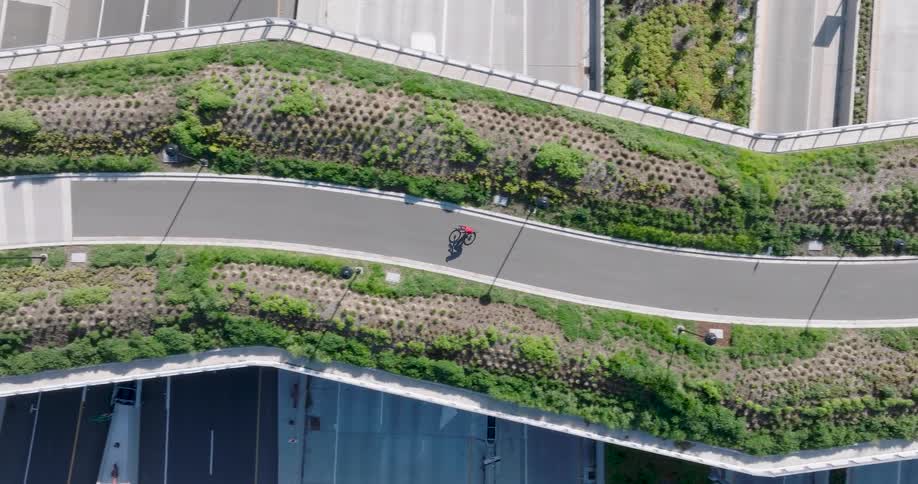 Top-down aerial footage follows a lone cyclist riding on a modern, green pedestrian and bike bridge crossing over a wide highway