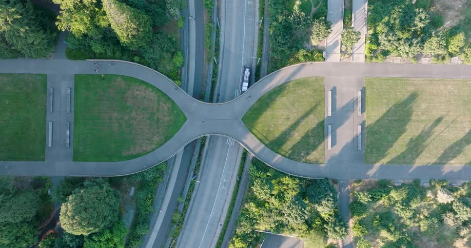 Top-down aerial view ascends over a unique hourglass-shaped pedestrian bridge and park above a city road with traffic