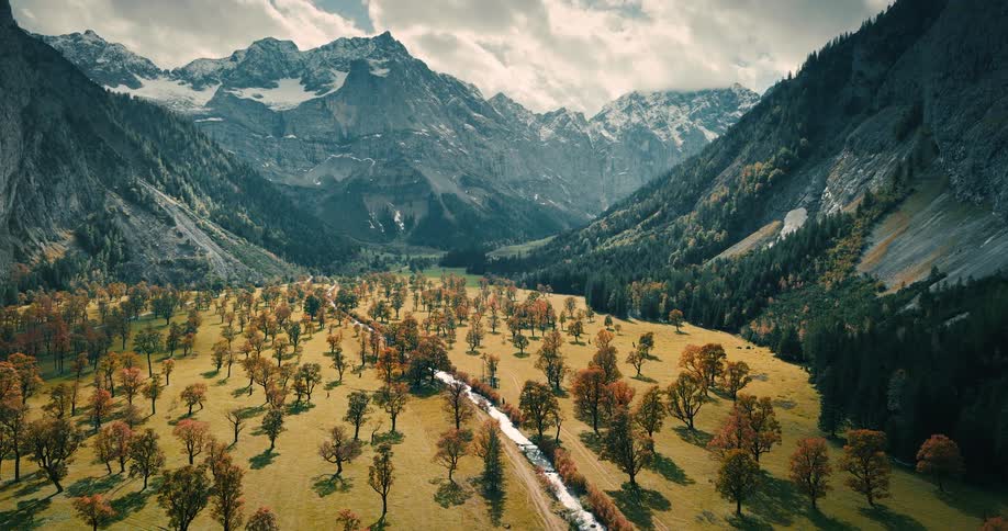 An idyllic alpine valley in autumn, with colorful trees, a flowing stream, and a backdrop of impressive snow-capped mountains