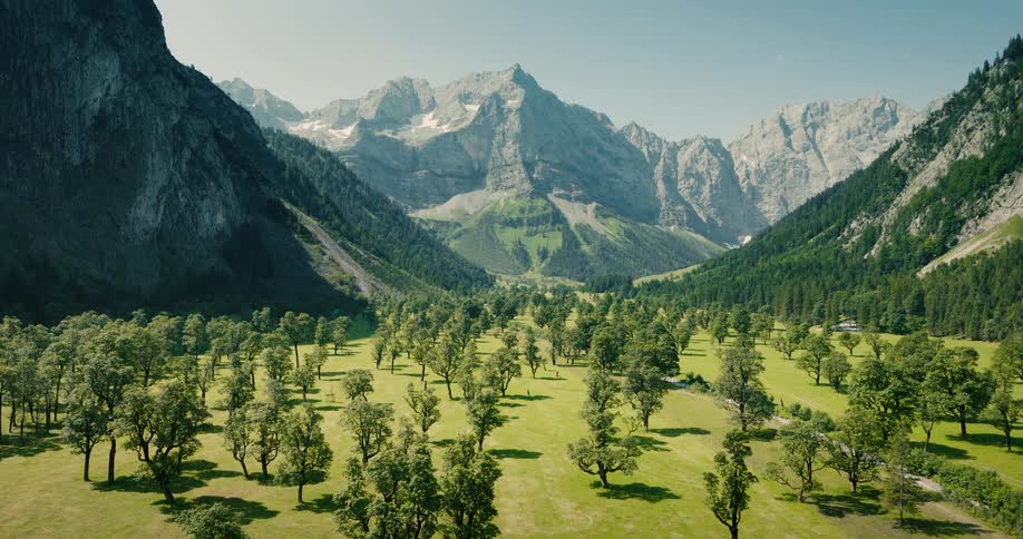 Breathtaking aerial view of a lush green valley with scattered maple trees nestled beneath the majestic, snow-capped Karwendel mountains in Austria