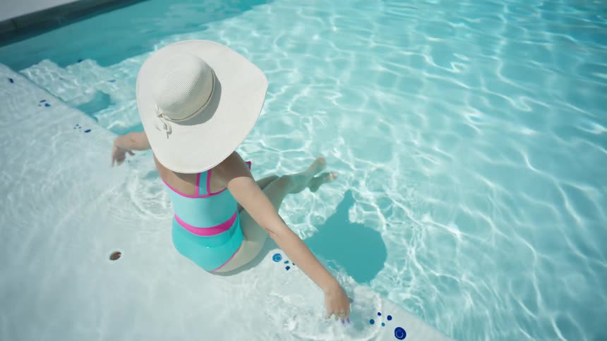 Stylish woman in a wide-brimmed hat and colorful swimsuit relaxes on the steps of a sparkling blue swimming pool on a sunny day