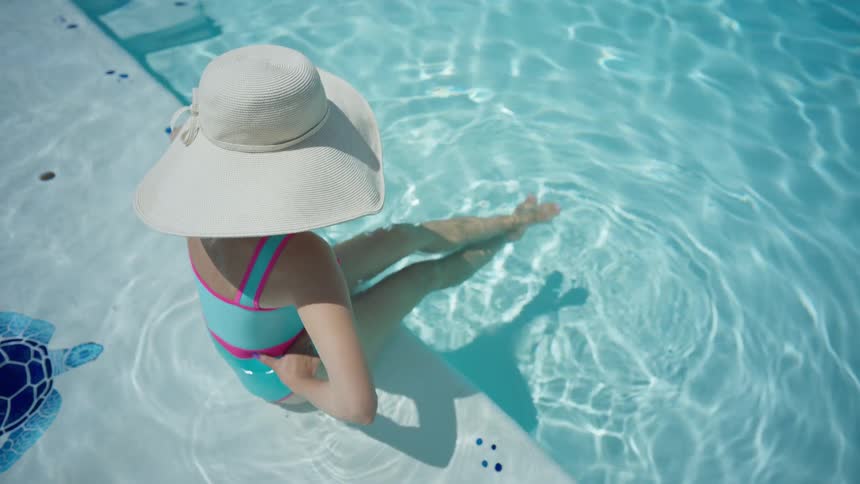 An overhead view of a girl in a large sun hat relaxing on the steps of a sparkling swimming pool on a sunny day