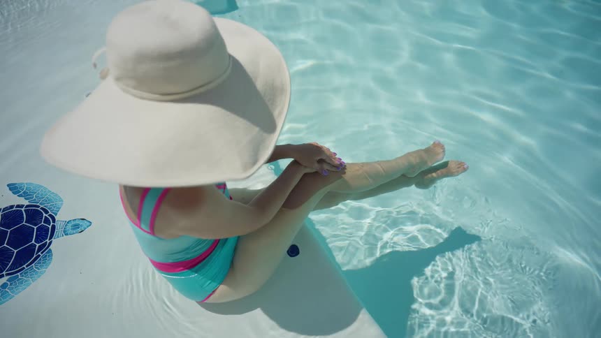 Elegant woman in a stylish white hat and colorful swimsuit enjoys a relaxing day in a sunlit swimming pool