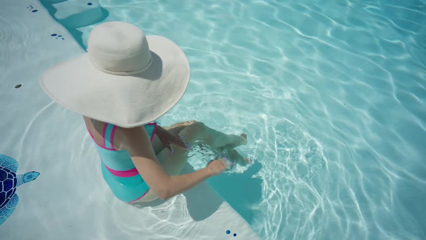 A high-angle view shows a woman in a colorful swimsuit and large white sun hat relaxing on the steps of a sparkling blue swimming pool
