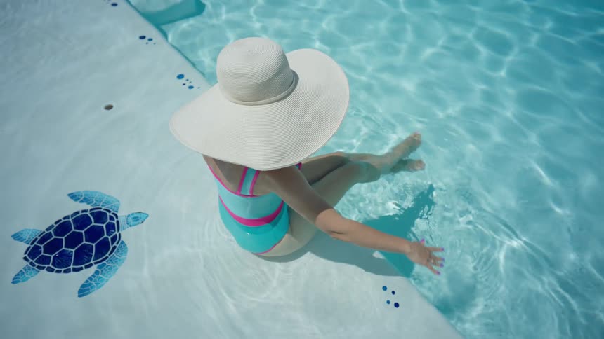 From a high angle, a woman in a wide-brimmed hat and colorful swimsuit enjoys the cool water while sitting on the steps of a sunny pool