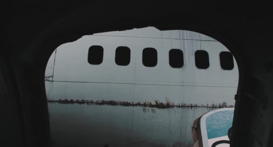 Eerie view of a rusty, abandoned airplane through a broken window frame in a vehicle graveyard