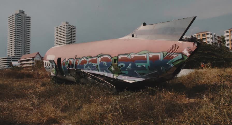 A derelict airplane fuselage covered in colorful graffiti sits abandoned in a grassy field with city skyscrapers in the background