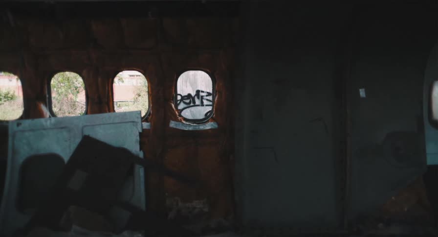 An eerie, static shot from inside the dark, dilapidated fuselage of an abandoned airplane with graffiti on the windows