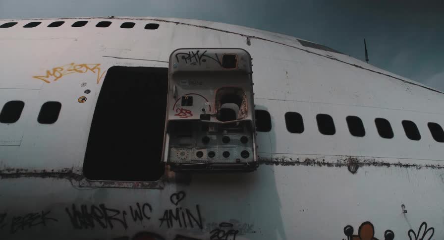 A low-angle view of an abandoned, graffiti-covered airplane fuselage with a broken open door against a gloomy sky