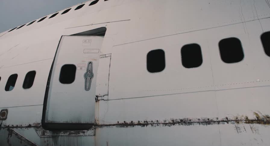 A static shot of a rusty, old, abandoned airplane fuselage with a slightly open door and windows on a cloudy day