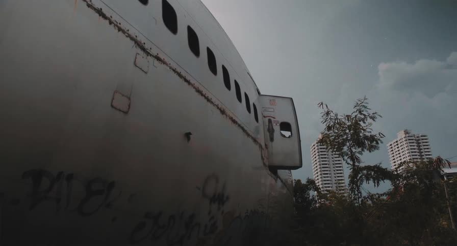 Low-angle time-lapse of a decaying, graffitied airplane in an overgrown lot, with city buildings visible under a cloudy sky