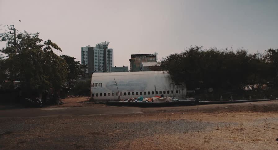 A wide static shot of an abandoned and graffitied airplane fuselage in a desolate lot with a modern city skyline in the background