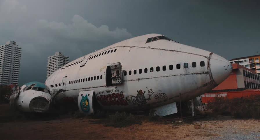 A graffiti-covered, abandoned jumbo jet rests in an urban airplane graveyard as storm clouds gather overhead