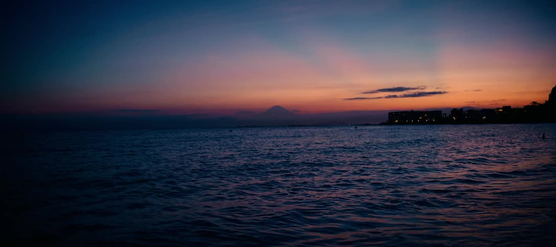 The silhouette of Mount Fuji stands on the horizon over a dark blue ocean as the sky glows with vibrant sunset colors